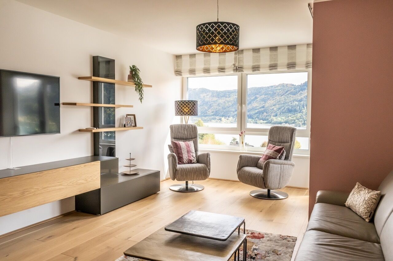 Una sala de estar moderna con una televisión de pantalla plana, estanterías de madera, dos sillones, un sofá, una mesa de centro y un amplio ventanal con vistas a un paisaje montañoso.