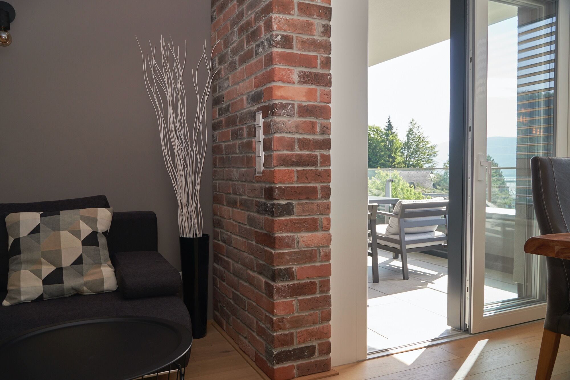 Un salon moderne avec un mur d’accent en briques, un canapé noir, un coussin à motifs et une porte en verre donnant sur une terrasse extérieure avec une table et des chaises.