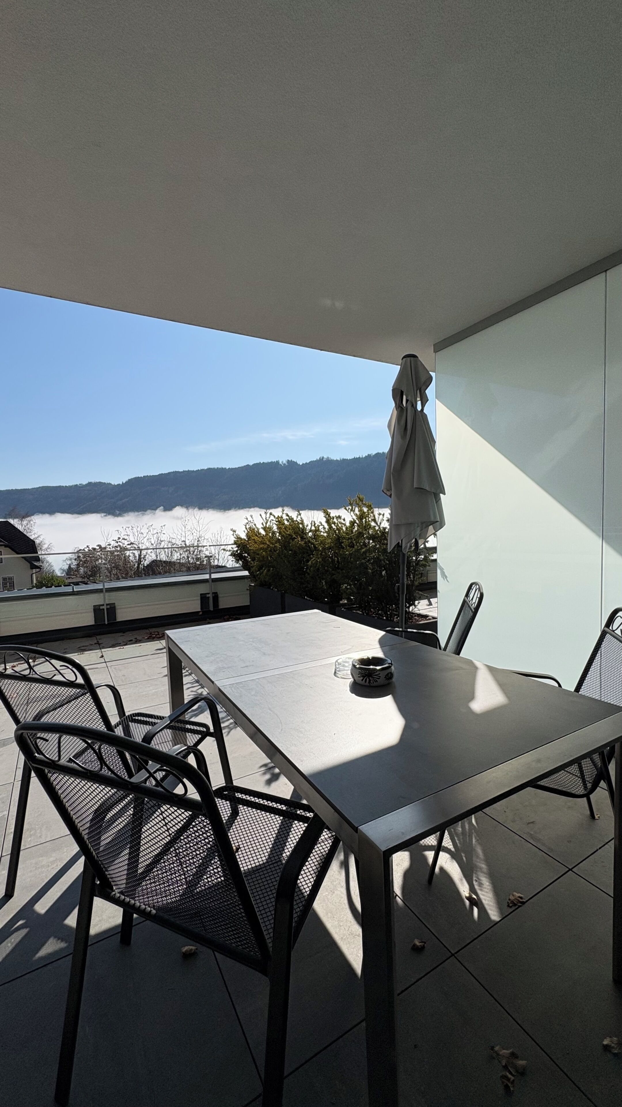 Un espacio moderno para comer al aire libre con mesa y sillas, y vistas a una cadena montañosa, además de una sombrilla.