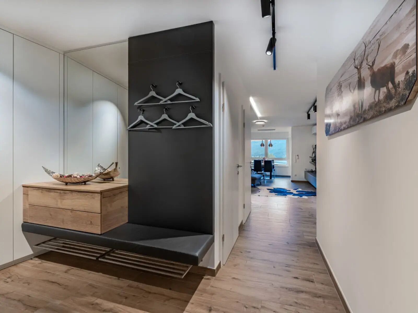 Een moderne hal met een houten bankje, een zwarte wand met hangende kleding en een groot schilderij aan de rechterkant.