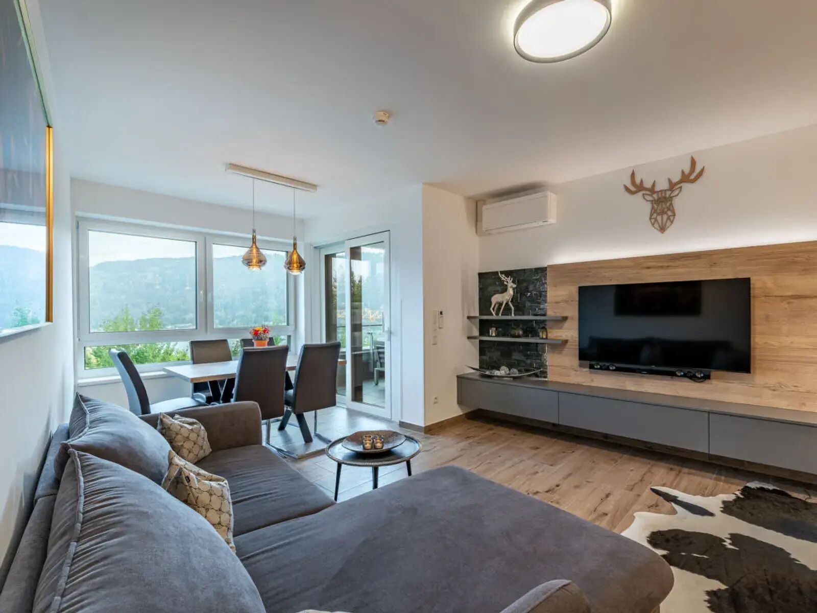 A modern living room with a large grey sofa, a flat-screen TV mounted on a wooden wall, and a dining area with a table and chairs.