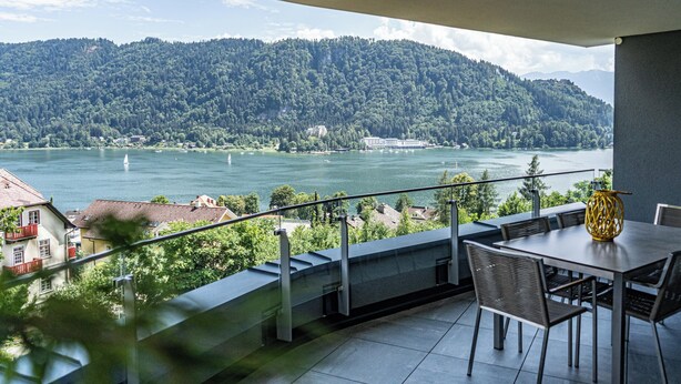 Ein Balkon mit Tisch und Stühlen, der auf einen See mit Booten und Gebäuden im Hintergrund blickt.