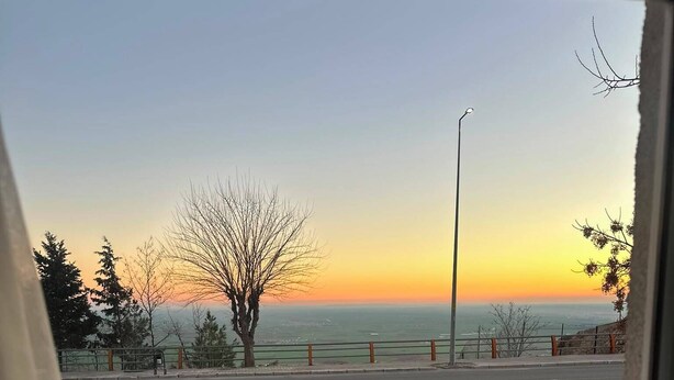 Ein Balkon mit Metallgeländer, Blick auf einen Sonnenuntergang über dem Meer und ein Baum im Vordergrund.