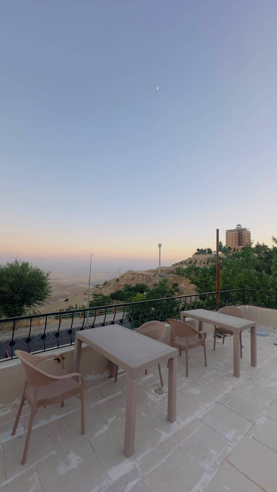 Una terraza con mesas y sillas, con vistas a un paisaje montañoso y un edificio distante.