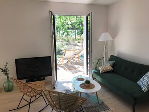 A living room with a sofa, a TV, a wicker chair, a small table, and a view of an outdoor patio with a chair and plants. - Maison 3 chambres au calme, jardin privatif et piscine partagée (La Teste-de-Buch)