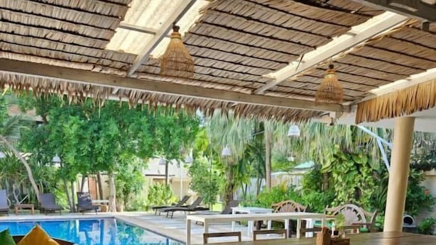 A poolside area with wooden furniture, a thatched roof, and lush greenery. - Fishermans Village Samui Yoga Retreat (Koh Samui)