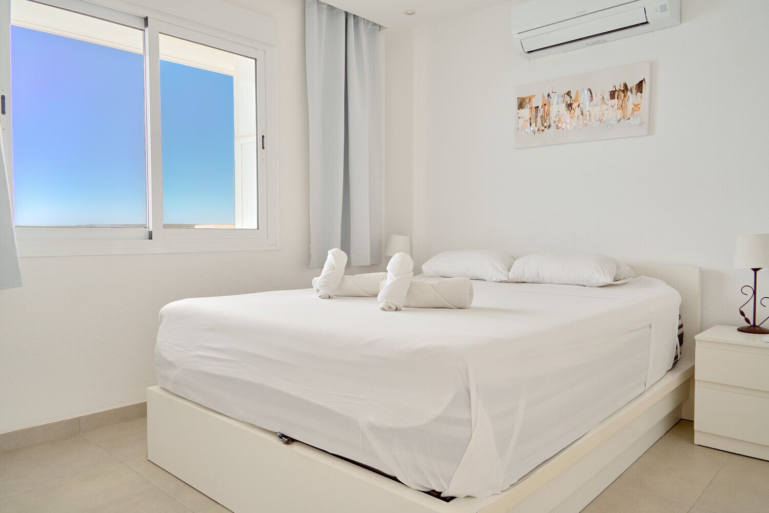 Une chambre à coucher blanche, avec un lit, une table de chevet, une lampe, une fenêtre et un climatiseur.
