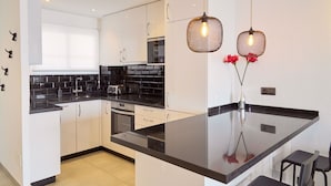 Een moderne keuken met witte kastjes, zwarte aanrechtbladen en een centraal eiland. Er hangen twee hanglampen met zichtbare lampen, en op het eiland staat een vaas met rode bloemen.