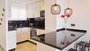 A modern kitchen with white cabinets, black countertops, and a central island. There are two pendant lights with exposed bulbs, and a vase with red flowers on the island. - Arce I 26 by Casasol (Nerja)