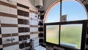 Une salle de bain avec un mur recouvert de carreaux à motifs, des toilettes, un bidet, un seau et une fenêtre donnant sur un paysage agréable.