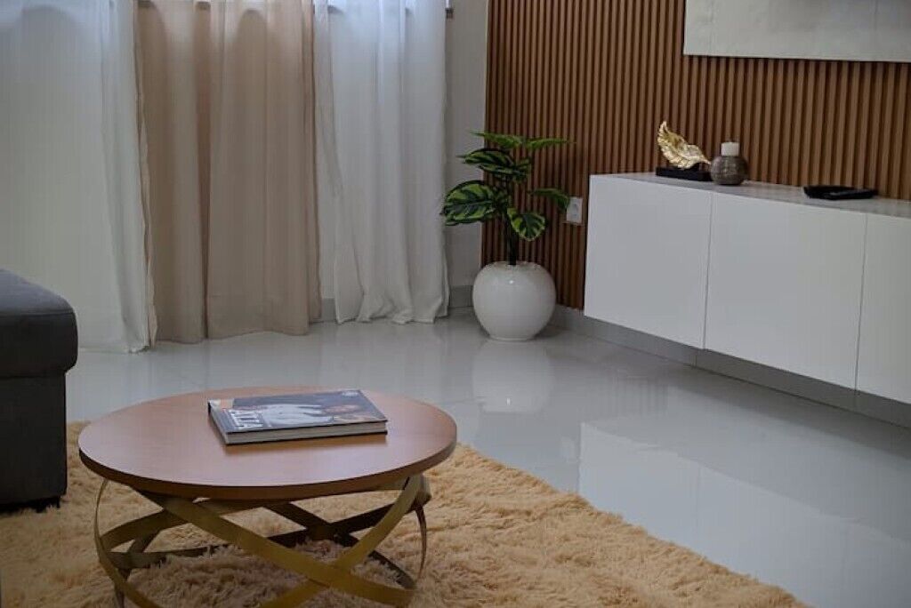 Ein modernes Wohnzimmer mit einem runden Holztisch, einer weißen Kommode, einer Topfpflanze und einem grauen Sofa.
