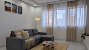 A modern living room with a sectional sofa, a round coffee table, and a large window with sheer curtains. - Modern & Stylish 1BR Apt | Cidadela | Ground Floor (Praia)