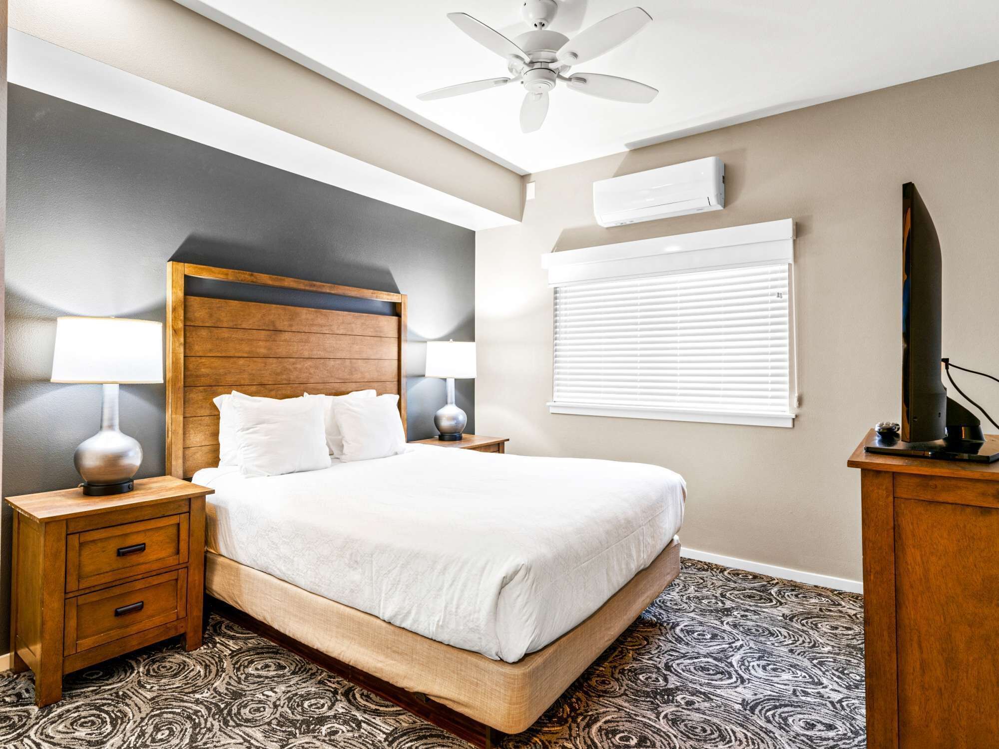 Quarto de hotel com cama, mesas de cabeceira, ventoinha no teto, ar condicionado e janela com persianas.