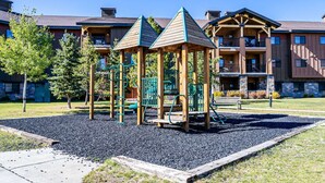 Ein Spielplatz mit Klettergerüsten und einer Bank, umgeben von Kies und Bäumen, mit mehrstöckigen Gebäuden im Hintergrund.