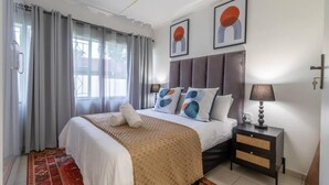 A bedroom with a bed, a nightstand, a lamp, and a window with curtains. - Greencroft Villa (Kempton Park)