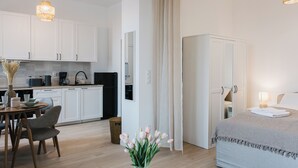 Une chambre d’hôtel moderne comprenant un lit, une table de chevet avec une lampe, une petite table avec un vase de fleurs, un coin cuisine avec des placards et des appareils électroménagers, et une salle de bain avec un rideau de douche.