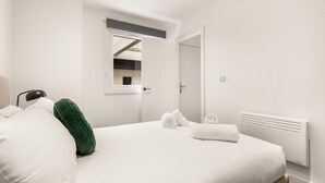 Et soveværelse med seng, sengetaske, grøn pude, sammenrullede håndklæder, et vindue og en radiator.