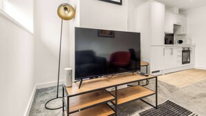 En moderne stue med et fladskærms-tv på et træstel, en gulvlampe og et køkkenområde med hvide skabe og hårde hvidevarer.