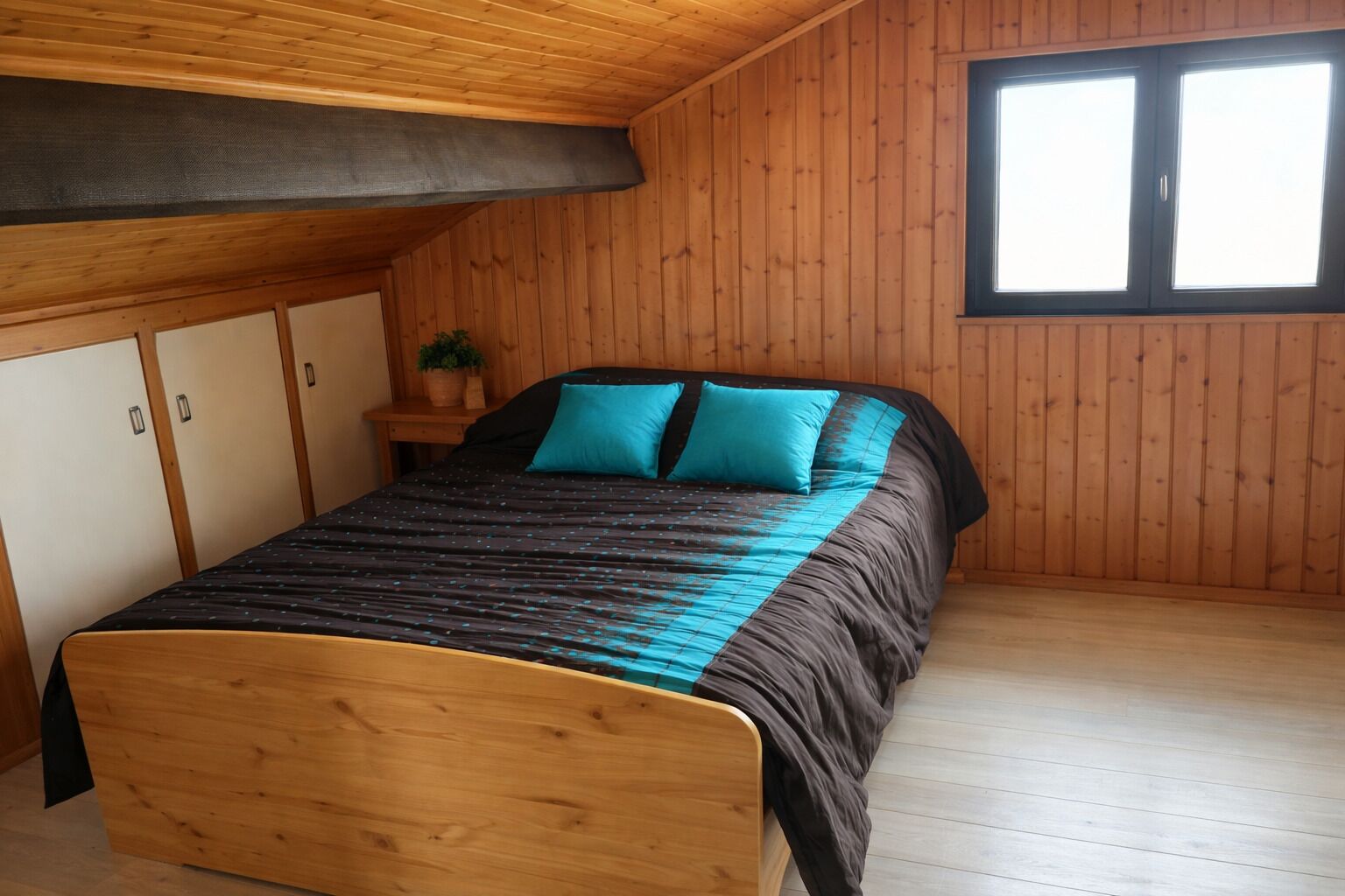 A wooden-paneled room with a bed, a window, and a small plant on a nightstand.