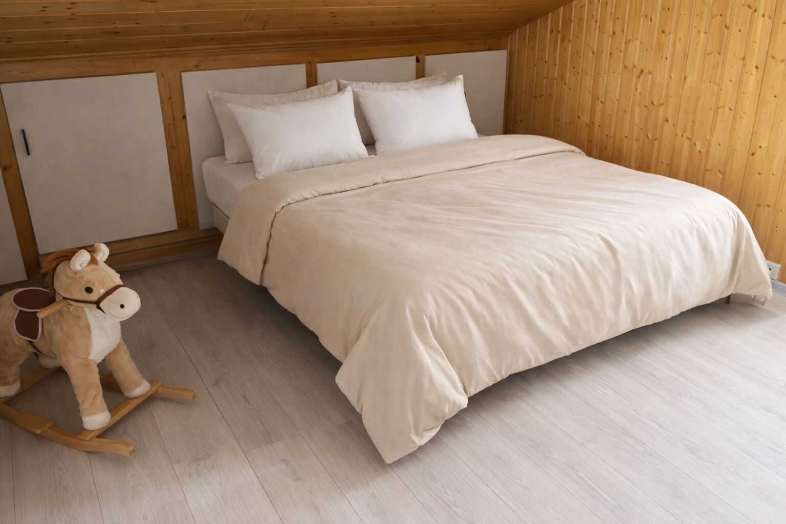 A bedroom with a wooden ceiling, a bed with a beige bedspread, two pillows, and a wooden rocking horse.