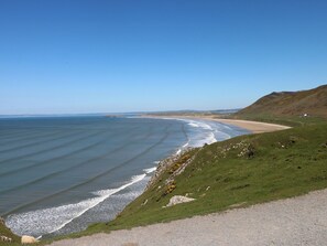 A coastal landscape with a beach, grassy hills, and a clear sky. - Butterslade (Swansea)