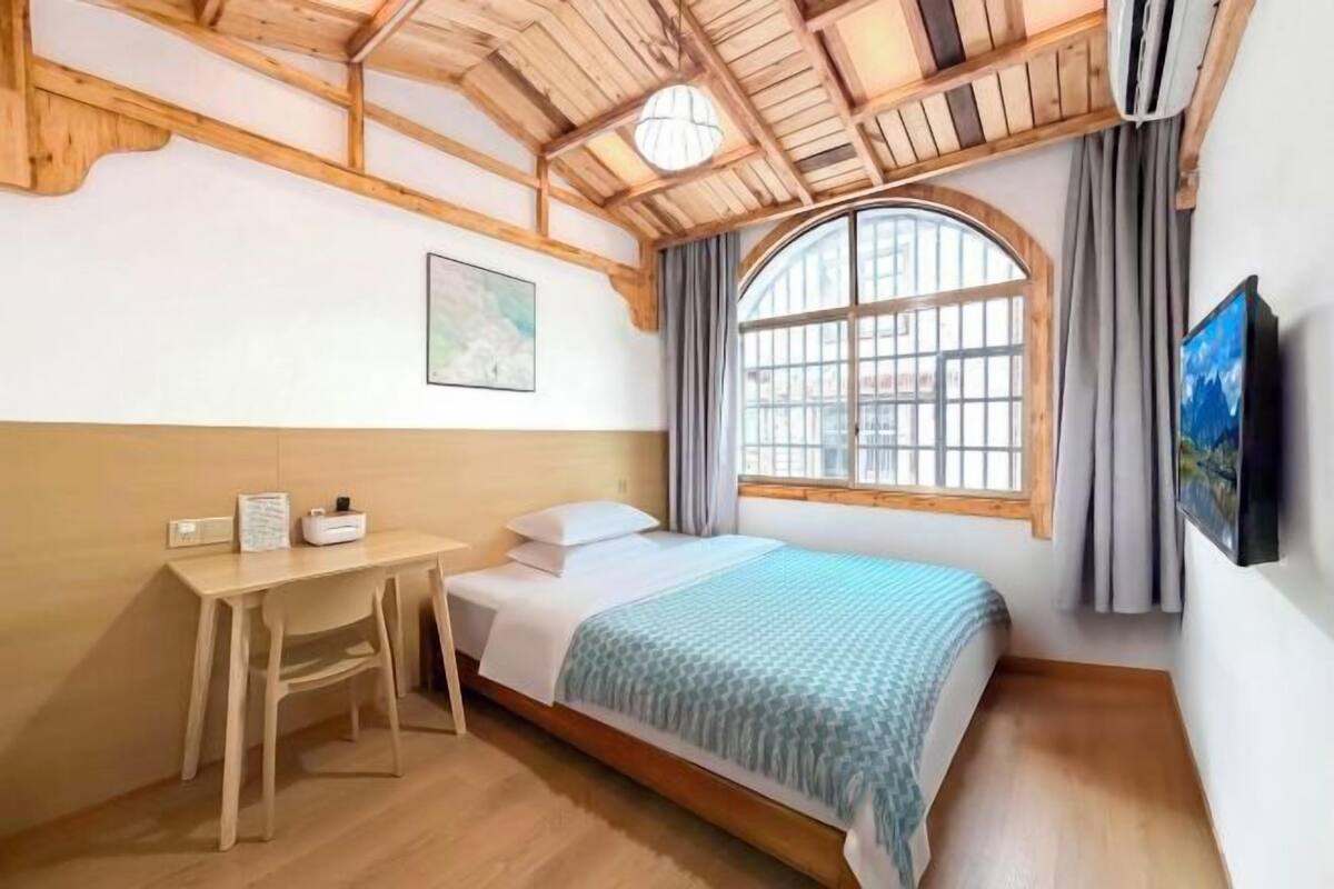 A bedroom with a bed, a desk, a chair, a window with curtains, and a painting on the wall.