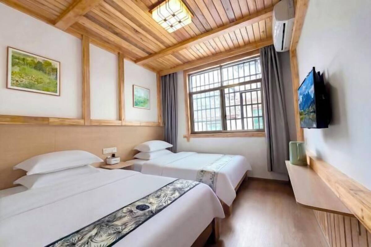 A hotel room with two beds, wooden ceiling beams, a large window with curtains, and a flat-screen TV mounted on the wall.