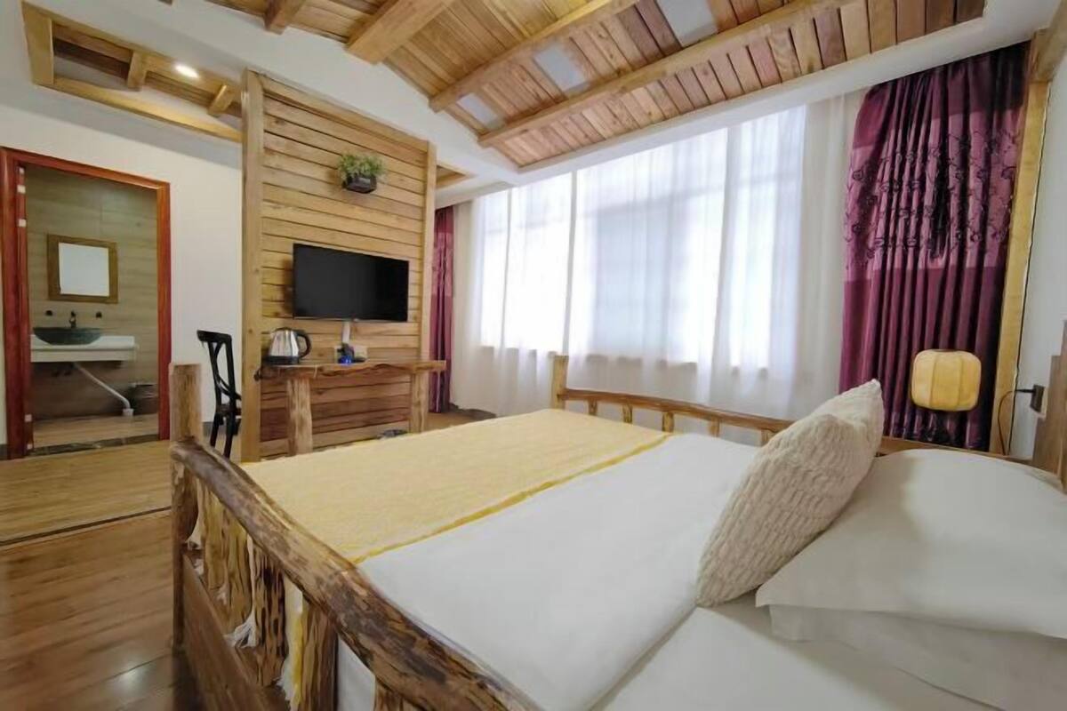 A bedroom with a wooden bed, a television on a wooden wall, a desk with a chair, and a window with curtains.