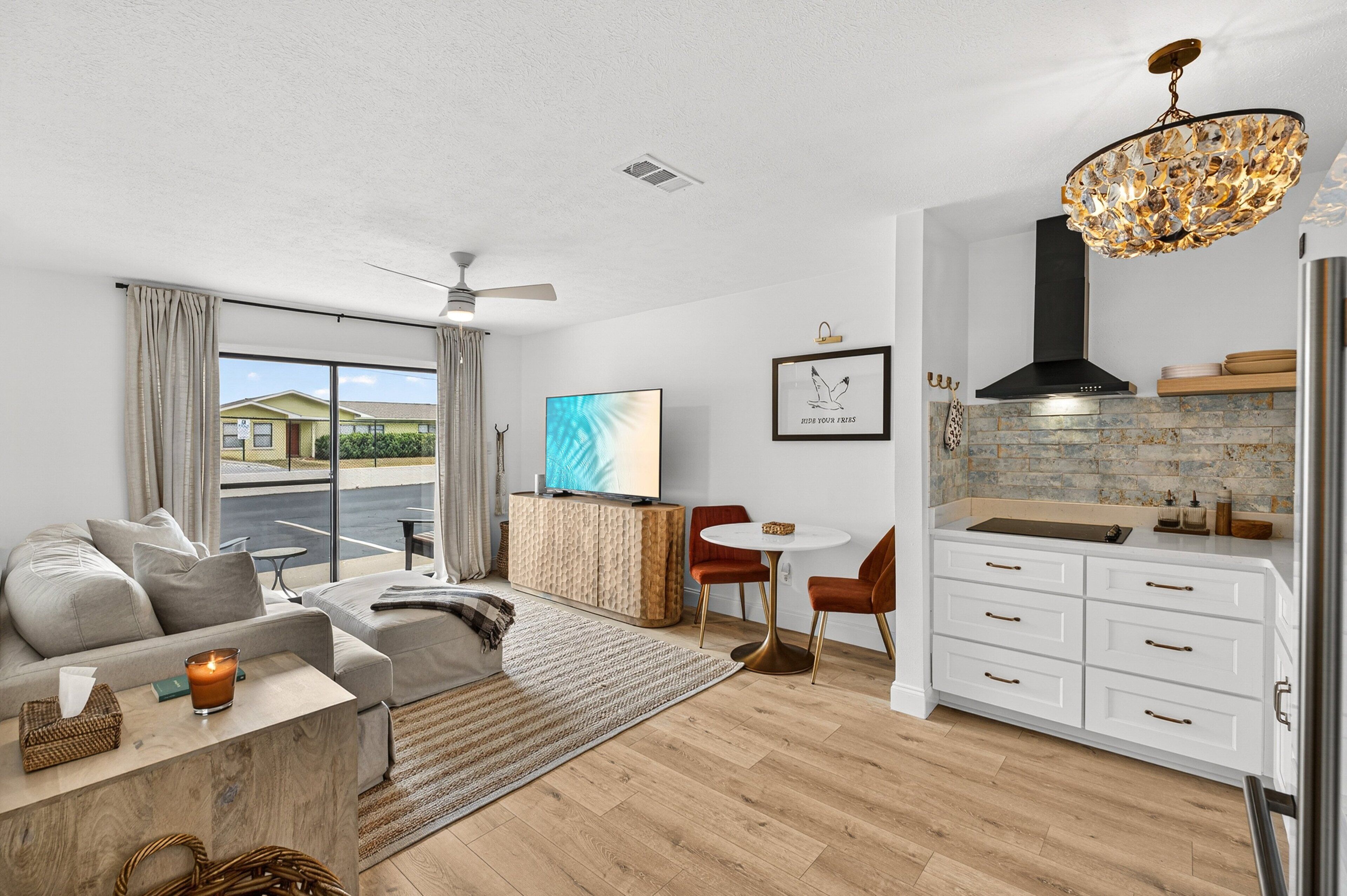 A modern living room with a sofa, coffee table, and a dining area with a table and chairs. There is a kitchen with white cabinets and a stainless steel refrigerator.