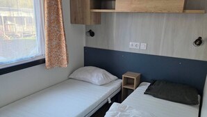 Ein gemütlicher Raum mit zwei Betten, einem Fenster mit Vorhängen, einem kleinen Holzregal und einer Wandleuchte.