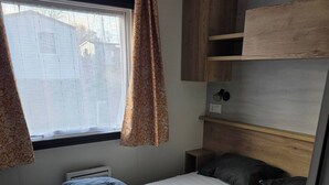 Ein kleines Schlafzimmer in einem Wohnmobil mit Bett, Kopfteil, Fenster mit Vorhängen und einem kleinen Regal.