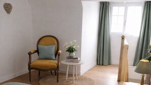 Un dormitorio con cama, una silla, una mesita con lámpara, una ventana con cortinas y una lámpara de pared.