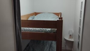 Una cama litera en un pasillo estrecho con una lámpara en el techo, una mesita de noche pequeña con lámpara, y un estante fijado a la pared.