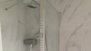 Ducha envuelta en vidrio con cabezal de ducha manual, grifo empotrado en la pared y un estante con un cabezal de ducha manual.