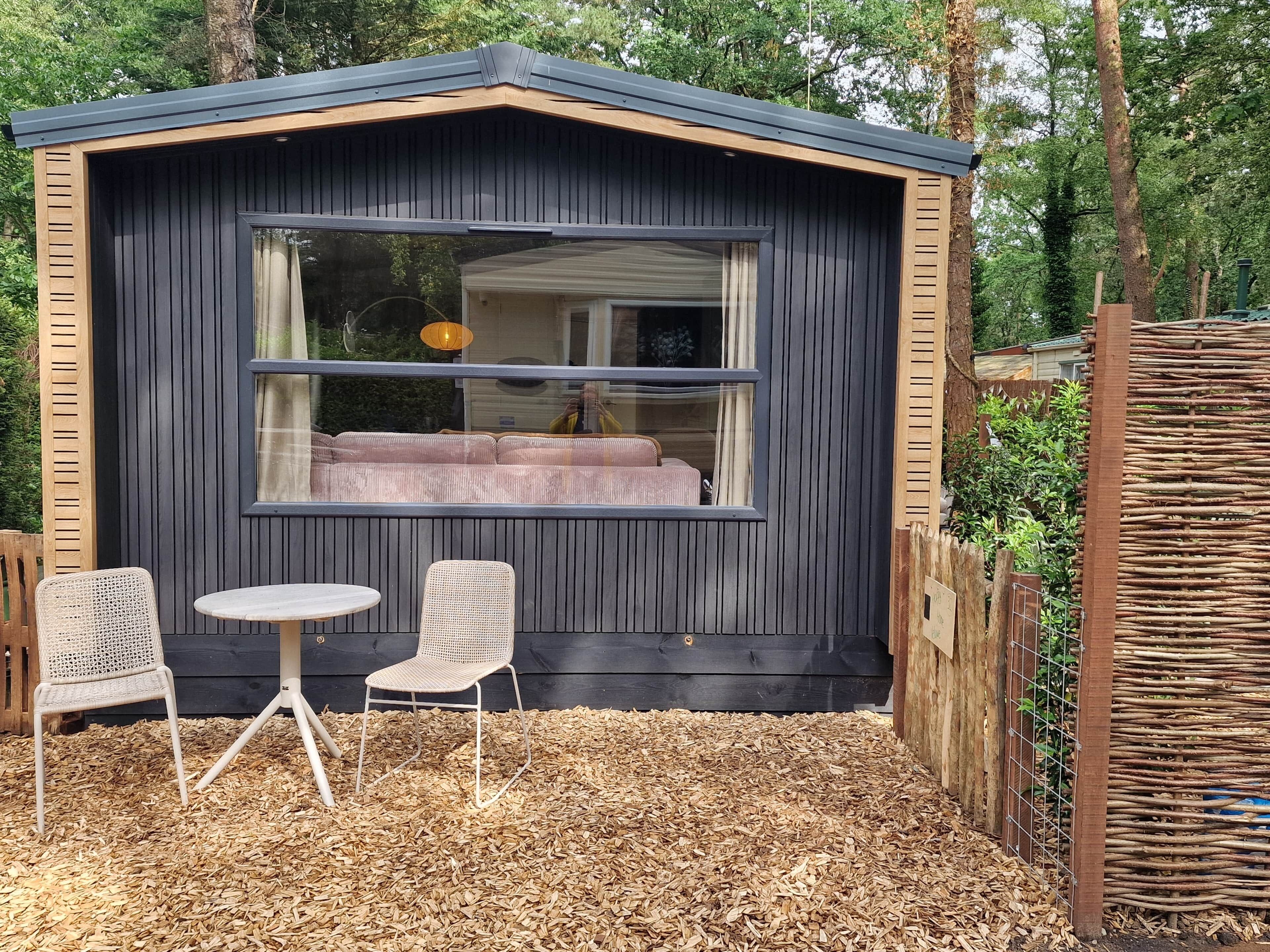 A small wooden cabin with a large window, two wicker chairs, a round table, and a sofa inside.