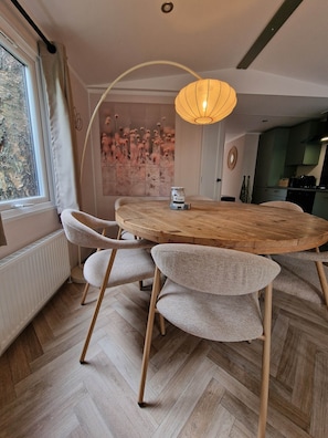 A dining area with a round wooden table, four woven chairs, a hanging light fixture, a radiator, and a view of the outdoors through a window.
