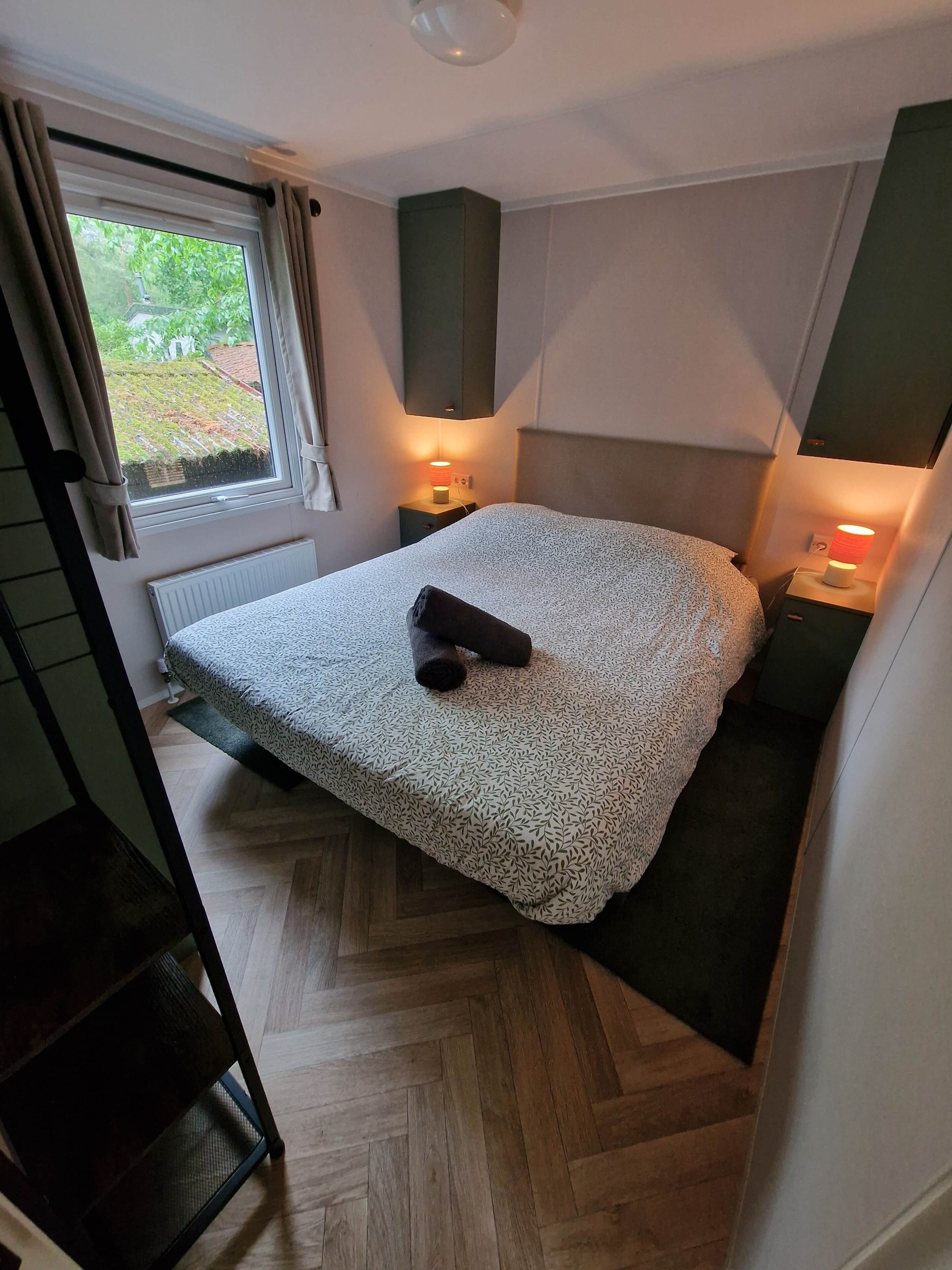 A compact bedroom with a bed, two bedside lamps, a window with curtains, and a radiator.