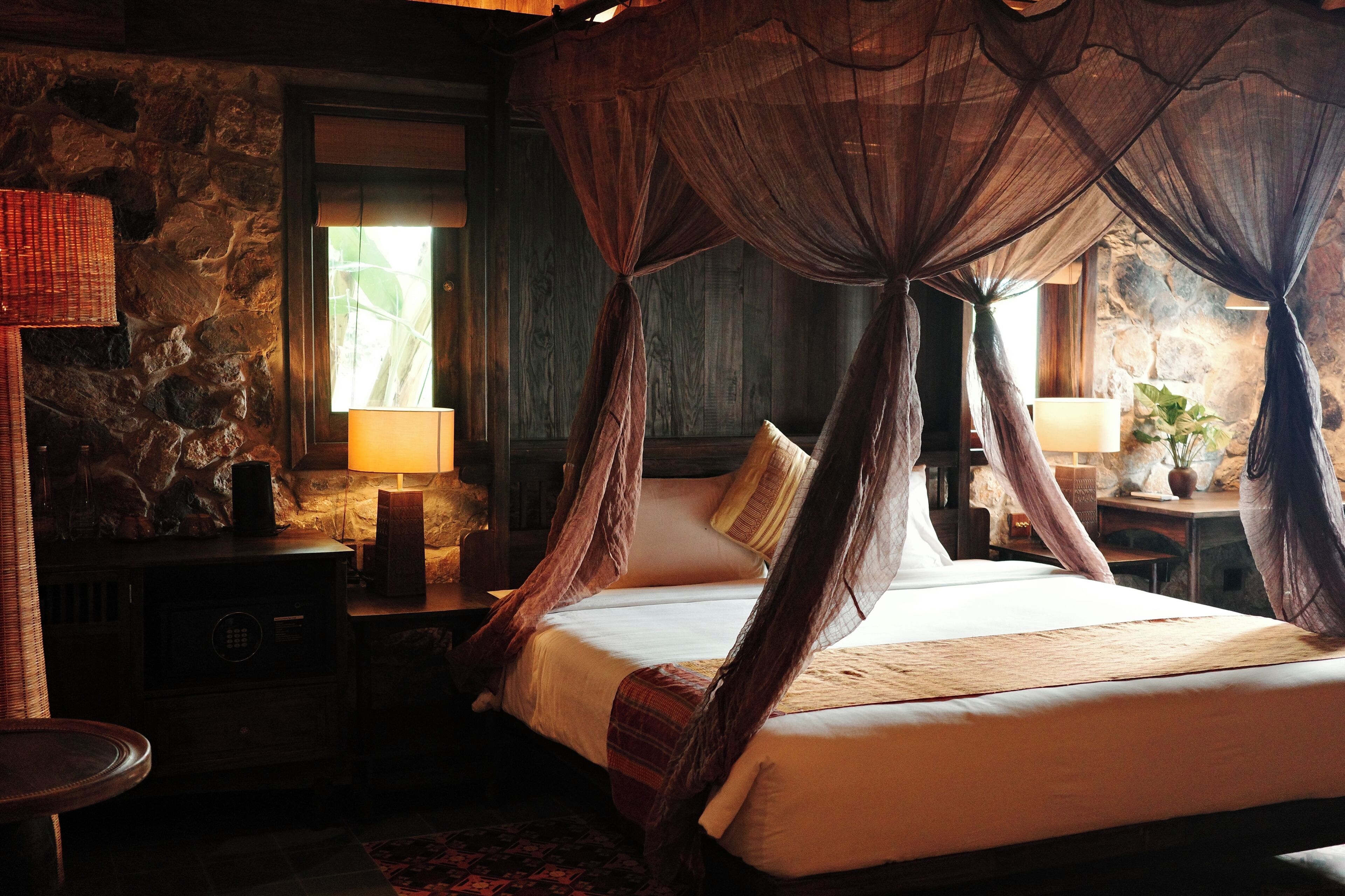 A bed with a canopy, a lamp, and a stone wall.
