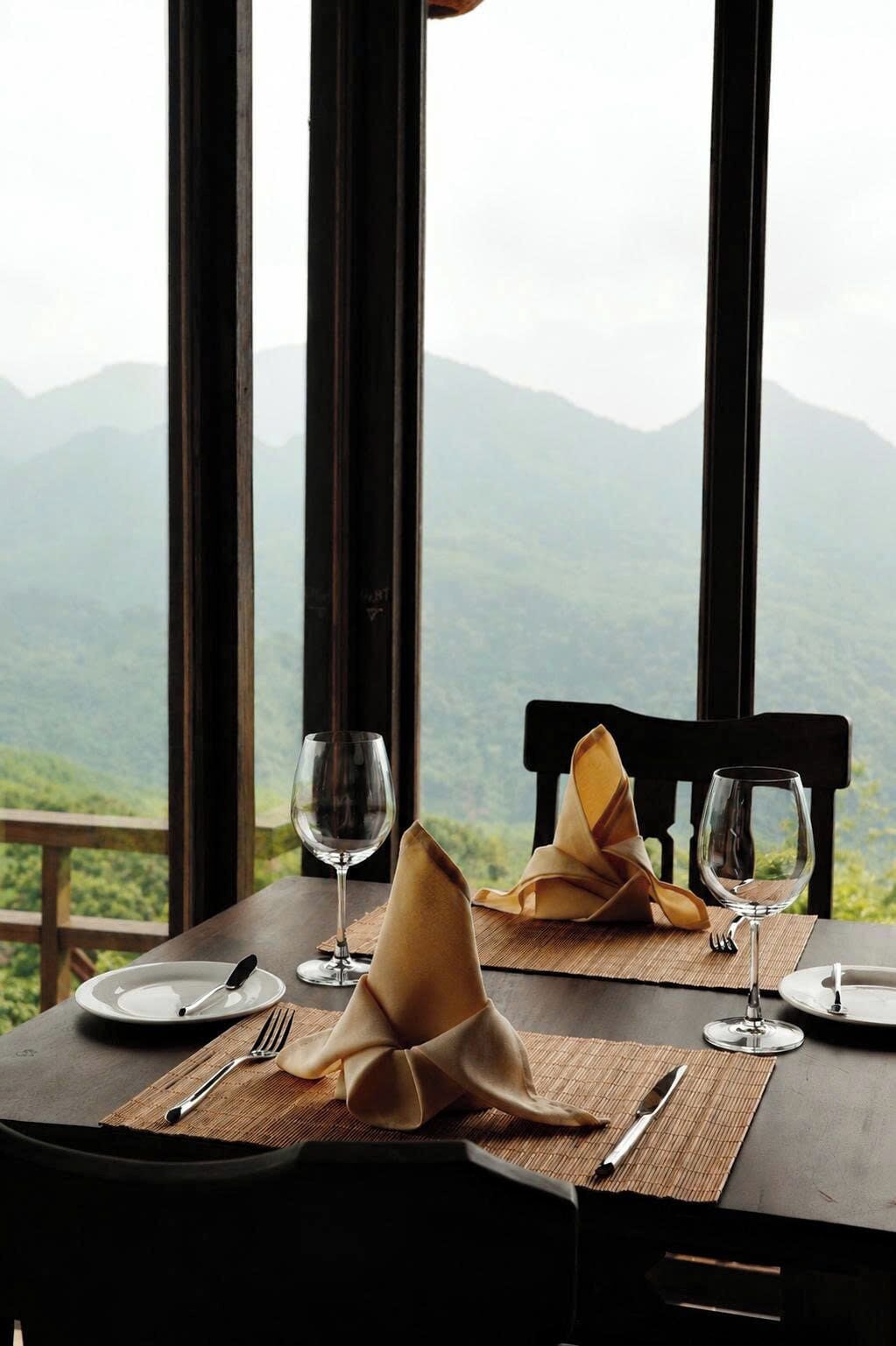 Mesa de jantar posta com pratos, talheres e guardanapos, com vista para montanhas através de grandes janelas.