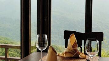 Mesa de jantar posta com pratos, talheres e guardanapos, com vista para montanhas através de grandes janelas.