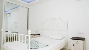 Uma cama de solteiro branca com cabeceira, mesa de cabeceira, pequeno armário e espelho.