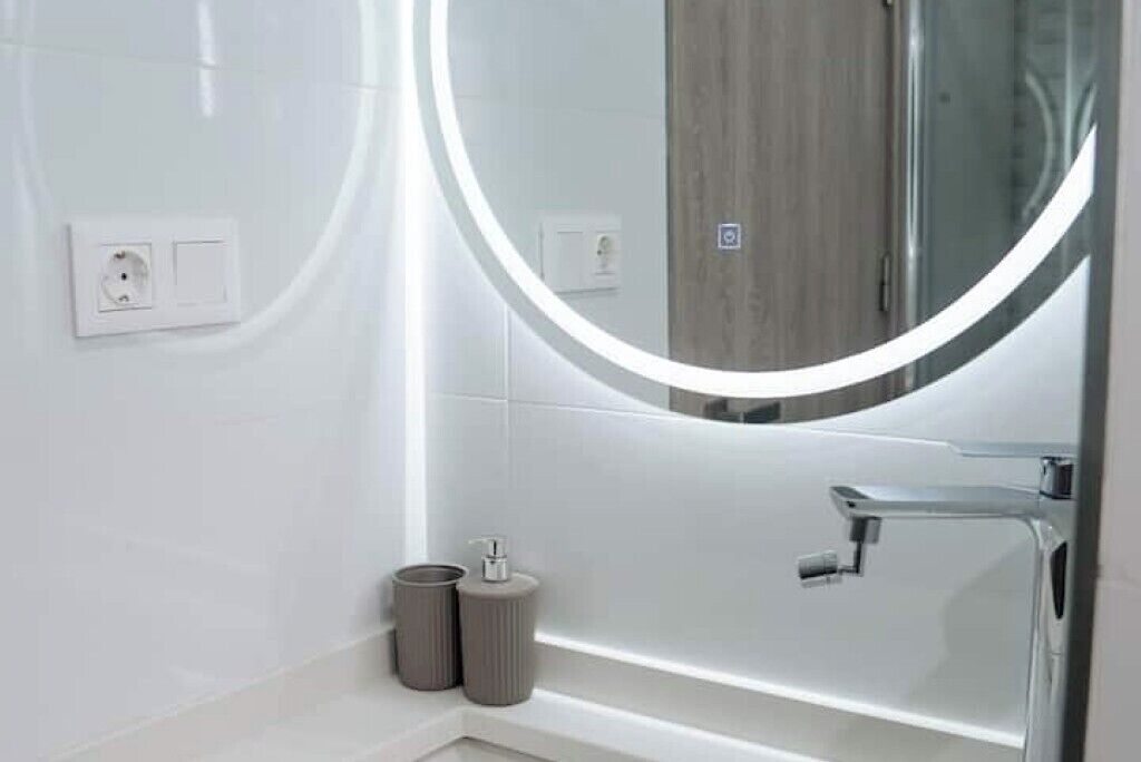Un bany de disseny modern, amb un mirall rodó amb llum, un lavabo blanc i un panell de fusta al fons.