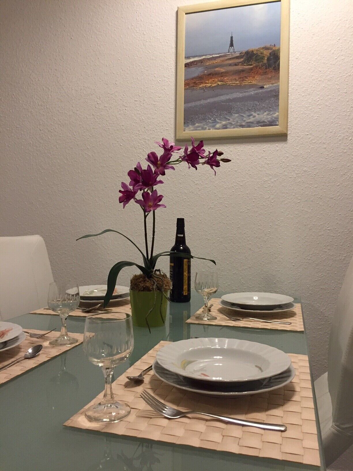 Een eettafel gedekt met borden, wijnglazen en een fles wijn, met een potplant en een lijst met foto aan de muur.