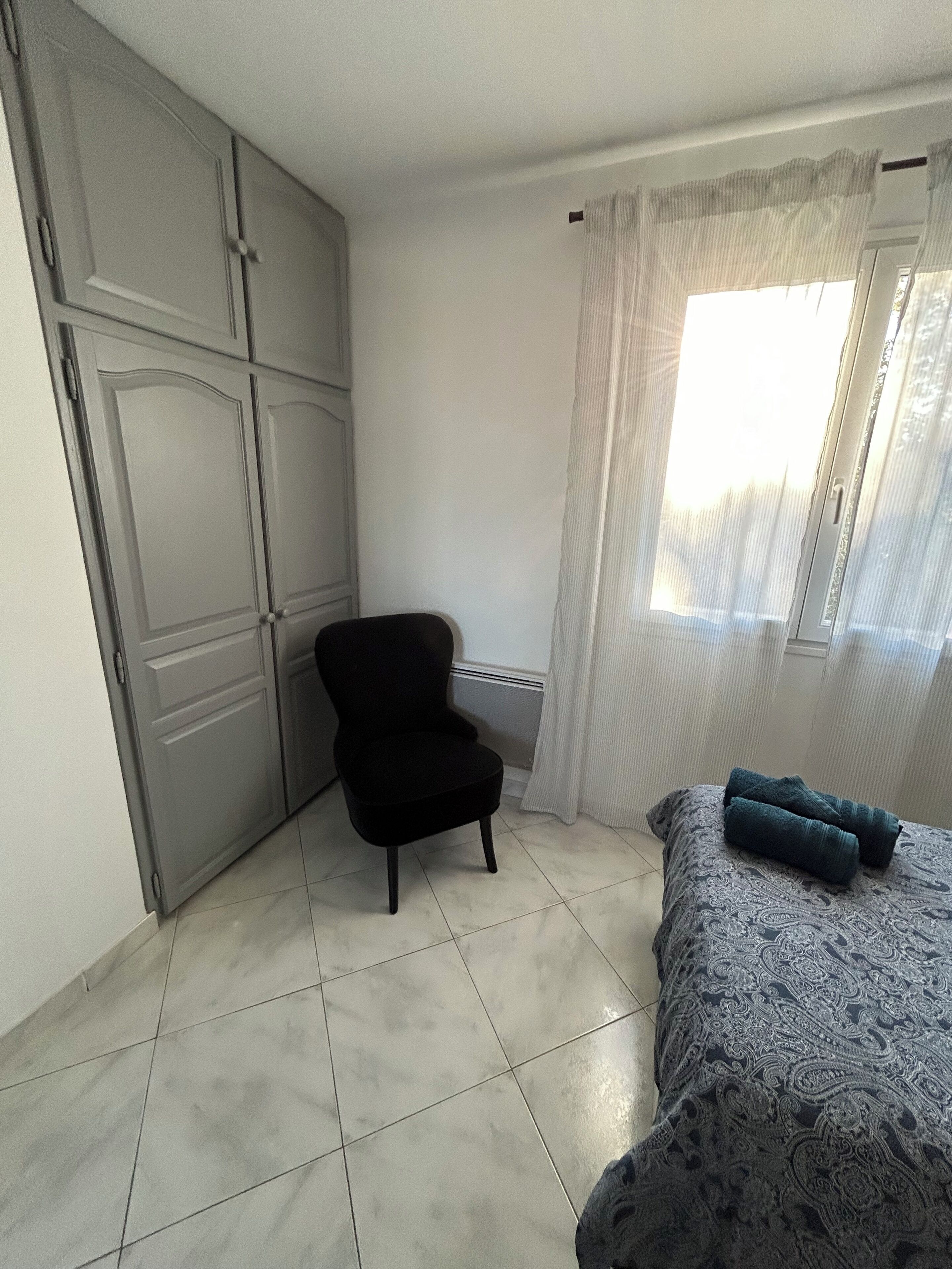 Un dormitorio con cama, una silla, un armario y una ventana con cortinas.