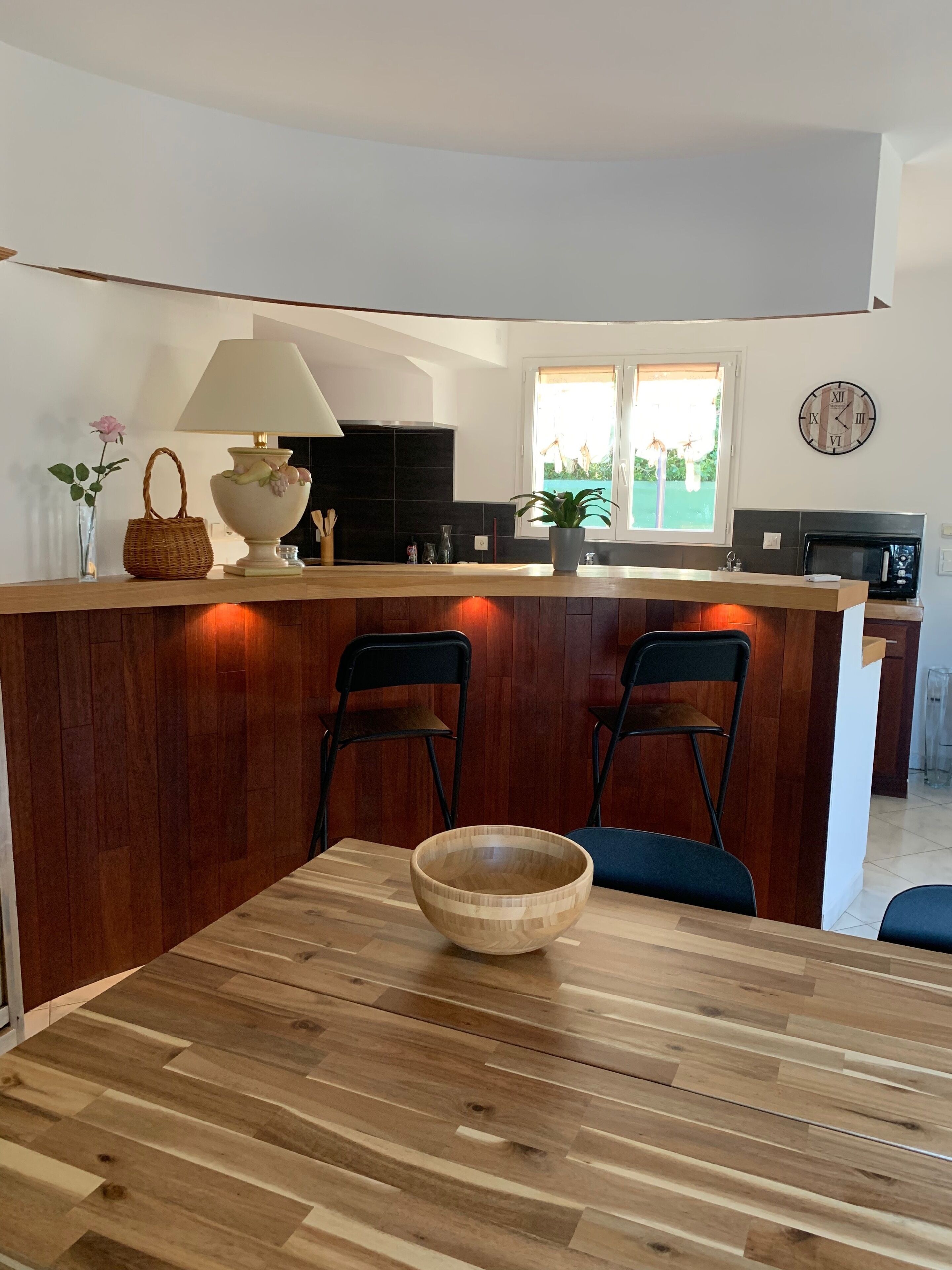 Een keuken met een houten kookeiland, twee zwarte stoelen, een houten eettafel met een kom, een lamp en een klok aan de muur.