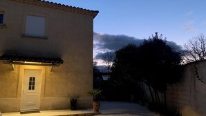 Un bâtiment avec un toit recouvert de tuiles, une porte blanche et une petite fenêtre. Devant la porte, on aperçoit un pot de fleurs, et un mur est partiellement recouvert de lierre.