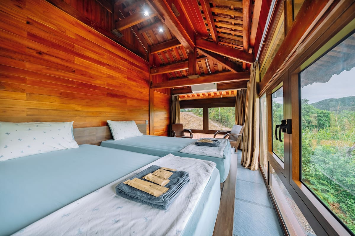 Una habitación con paredes y techo de madera, dos camas con ropa de cama azul, una ventana que da a vistas de follaje y un ventilador de techo.