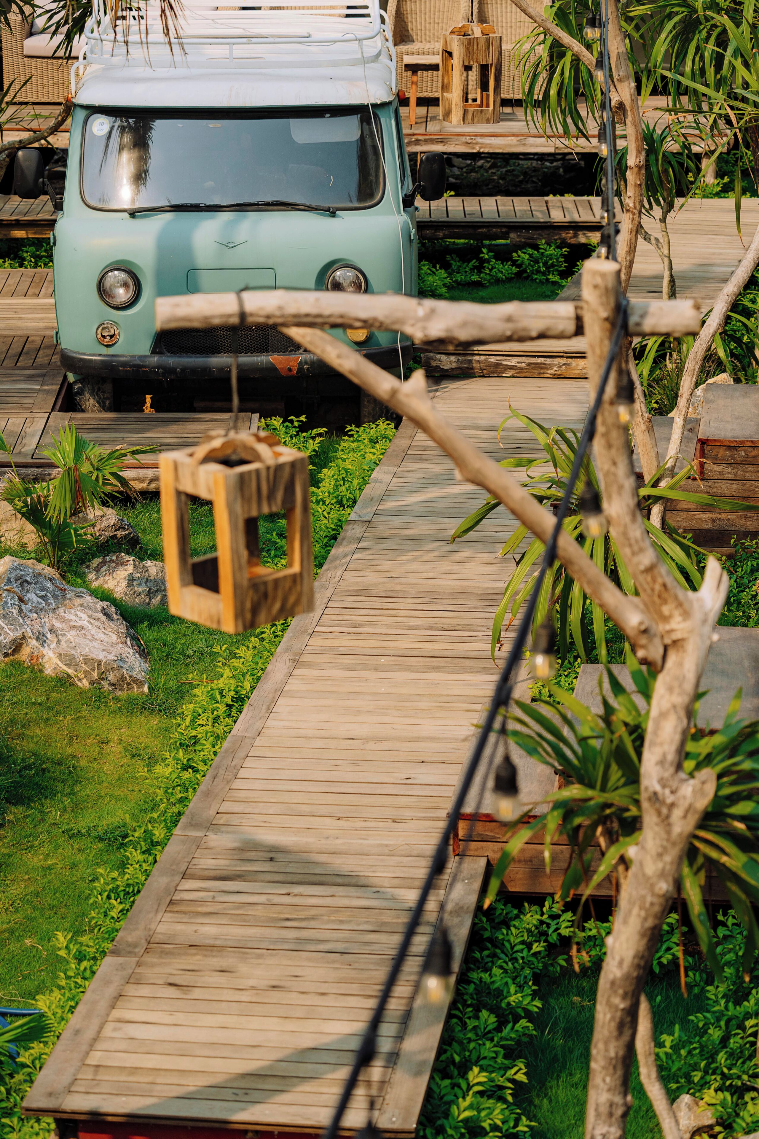 A wooden deck with a hanging lantern, surrounded by greenery and a vintage vehicle.