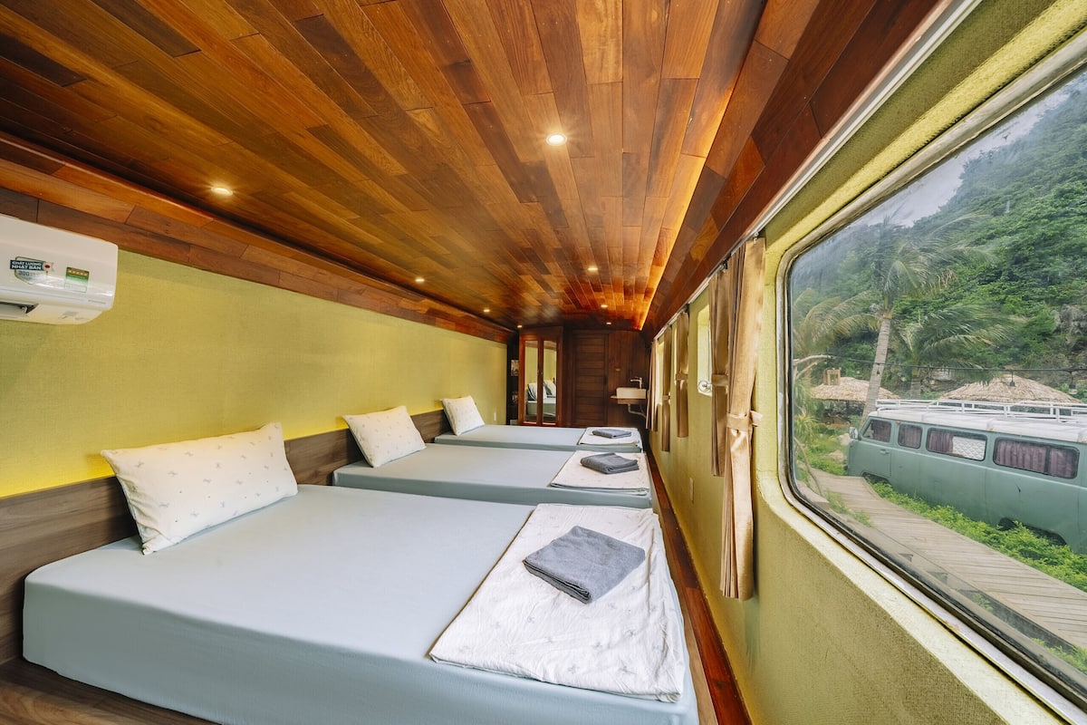 Un compartimento de tren con techos de madera, dos camas con almohadas, una ventana con vistas a follaje, y un lavamanos pequeño.