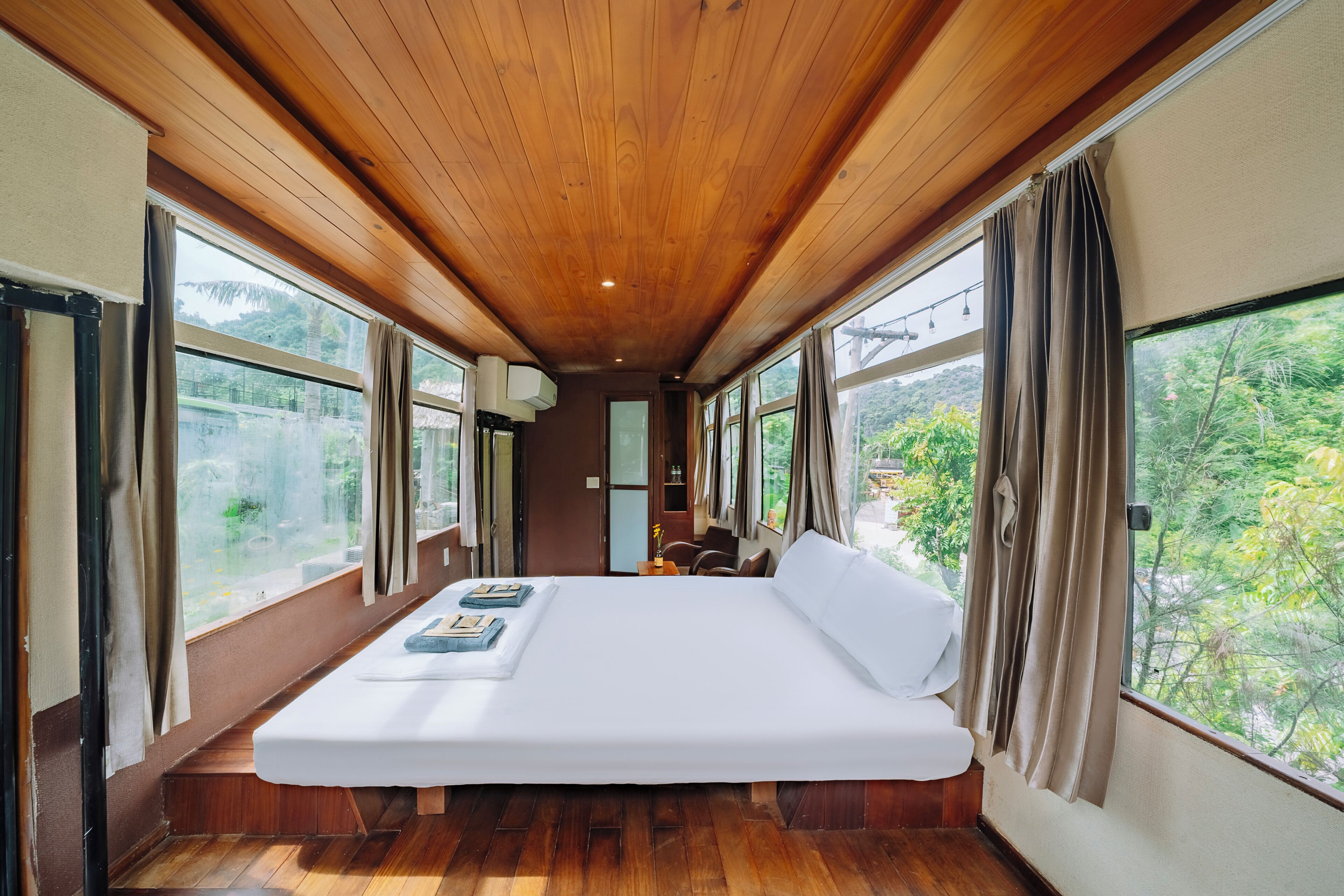 A room with a wooden ceiling, large windows, a bed, and a view of greenery outside.